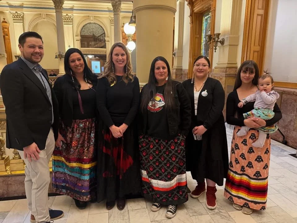 Seven people standing together inside a large, ornate building with tall columns, elaborate moldings, and warm lighting. Six women and one man, with one woman holding a young girl. The women are wearing colorful skirts or dresses, and the man is dres