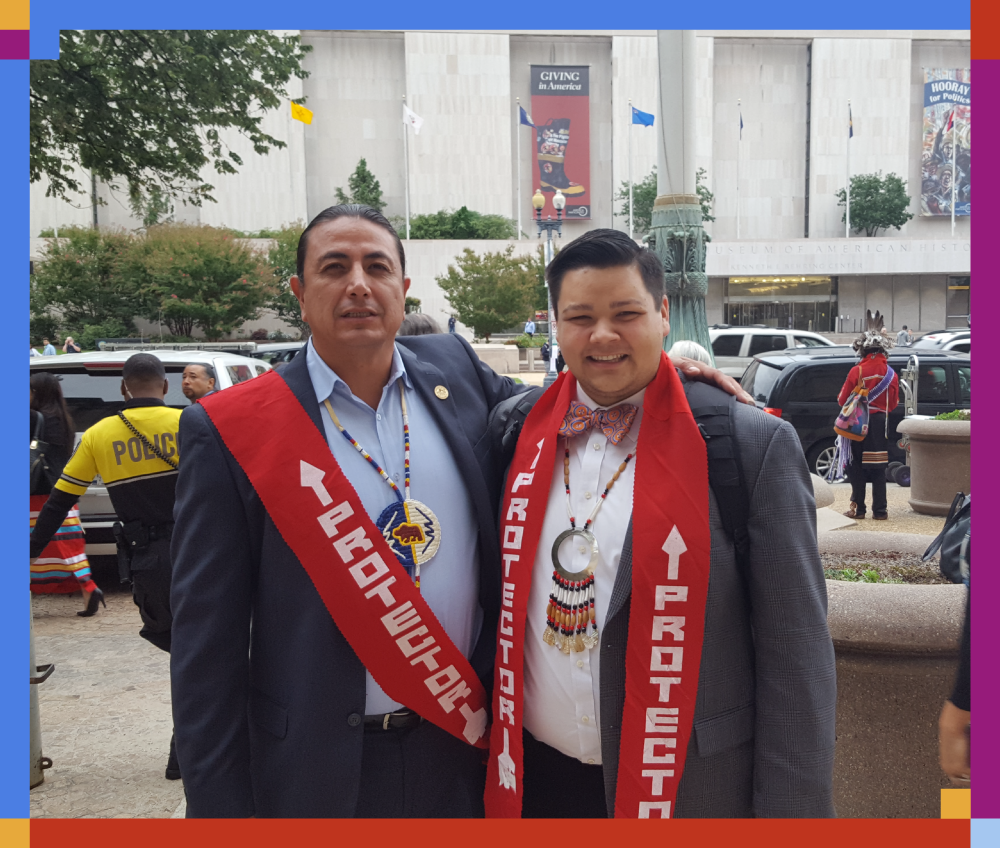 Two men in suits wearing red sashes with white text and beads around their necks, standing outdoors in front of a building, with people and police visible in the background. One man is smiling, and the setting appears to be a cultural or community event.