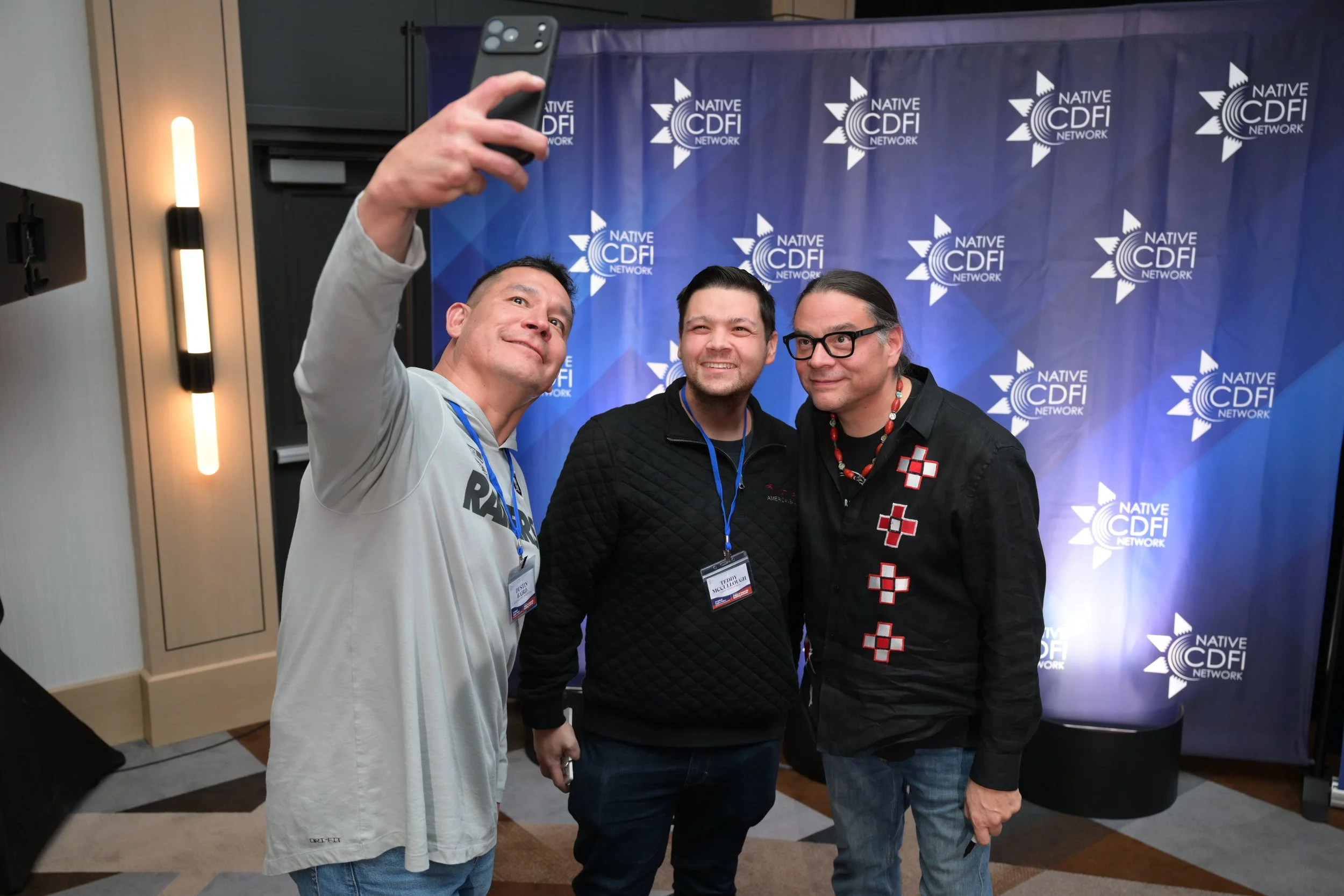 Three men taking a selfie in front of a blue backdrop with the 'Native CDF Network' logo, at a conference or event.