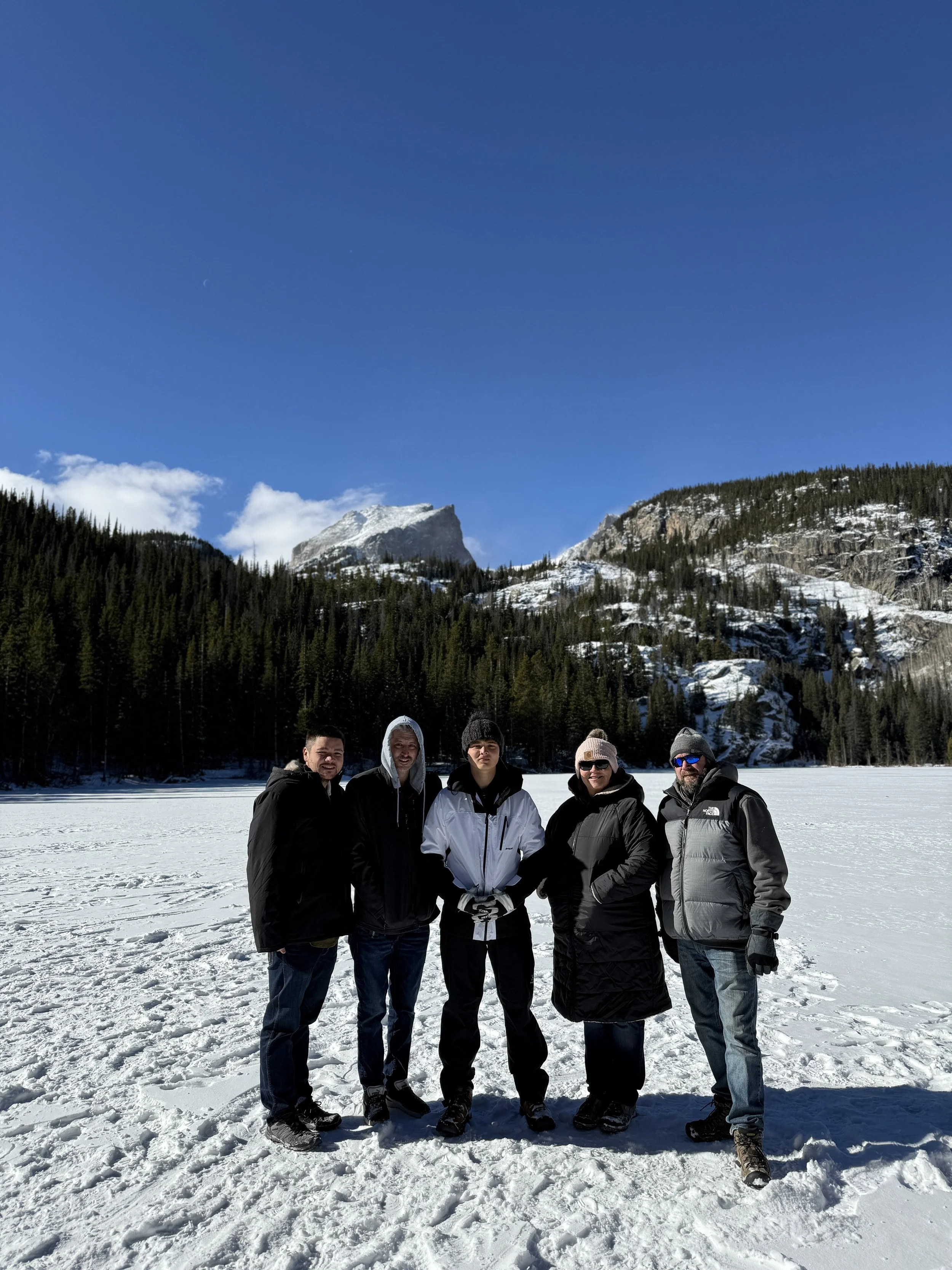 Five people dressed in winter clothing standing on snow-covered ground in a mountain and forest landscape with snow-capped peaks and a clear blue sky.