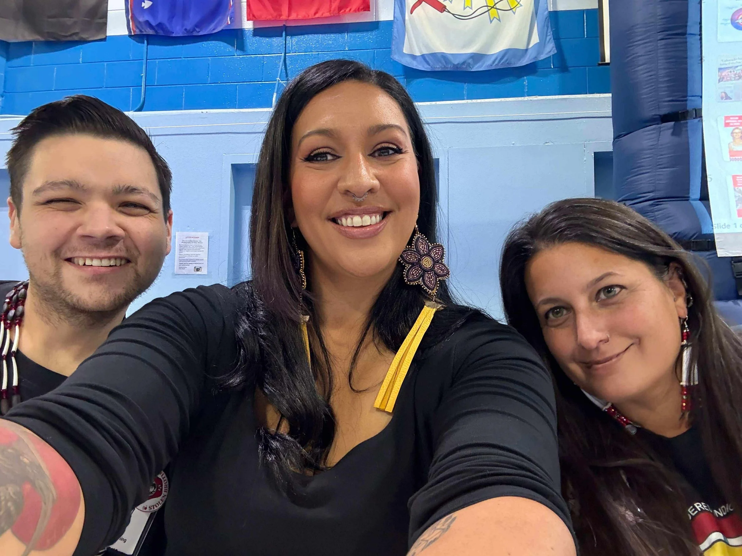 Three smiling people taking a selfie in front of a blue wall with banners and a poster.
