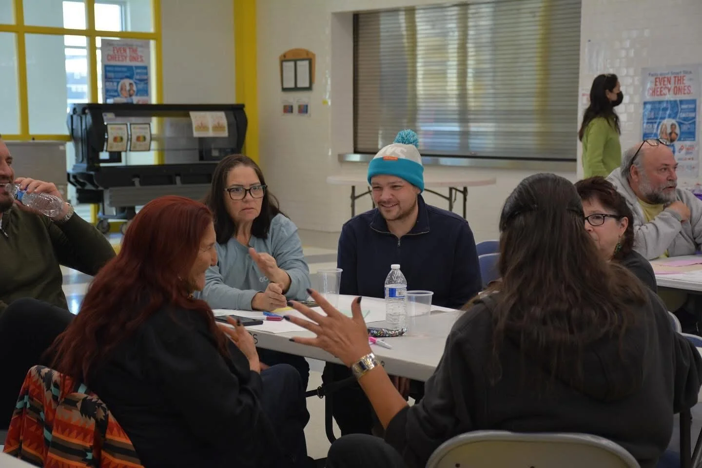 People sitting around a table in a group discussion indoors. Some are talking, one person is drinking water, and others are engaging in conversation.