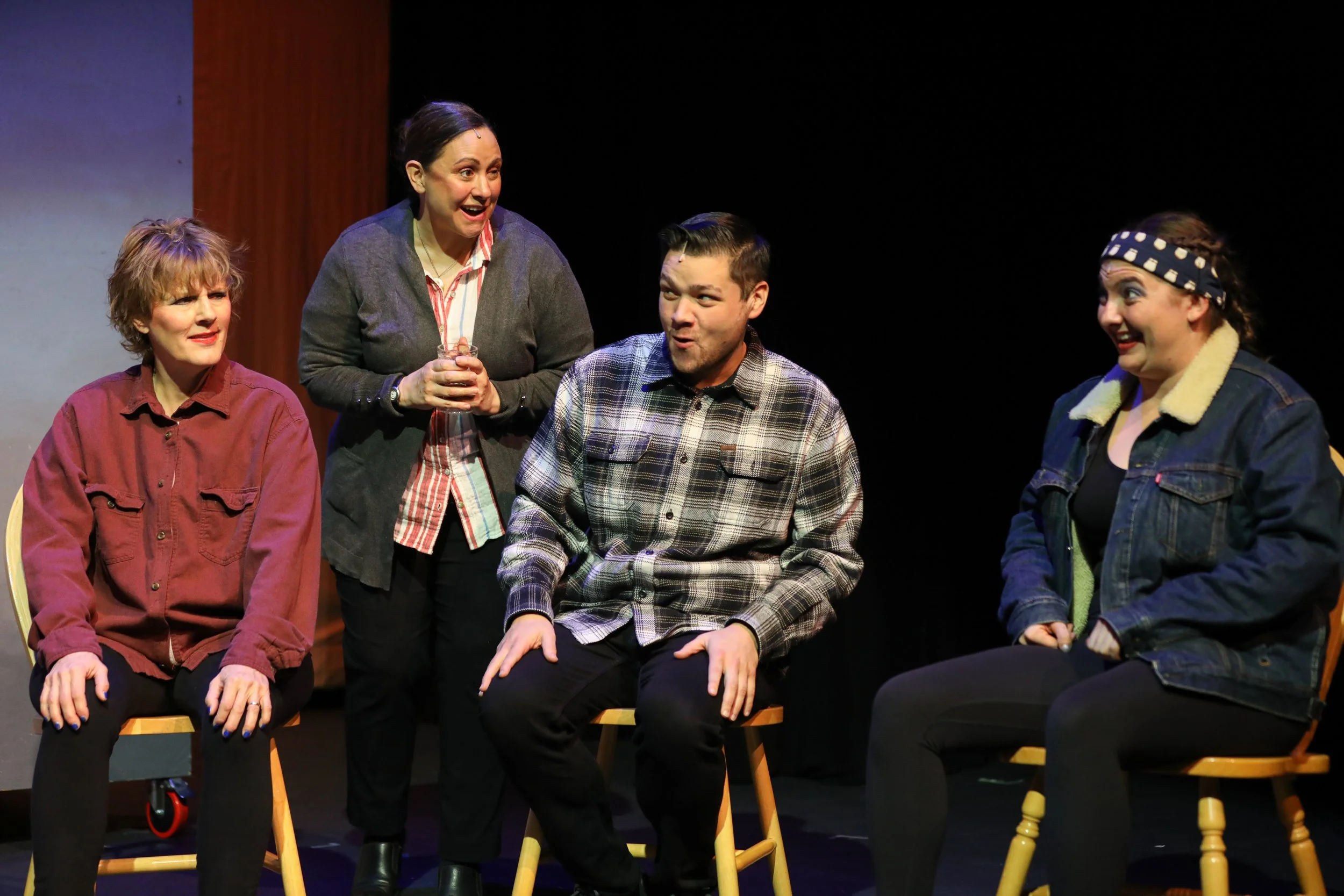 Four people performing a scene on stage, with three seated women and one standing woman engaging with them, all in casual, vintage-style clothing, with a dark background.