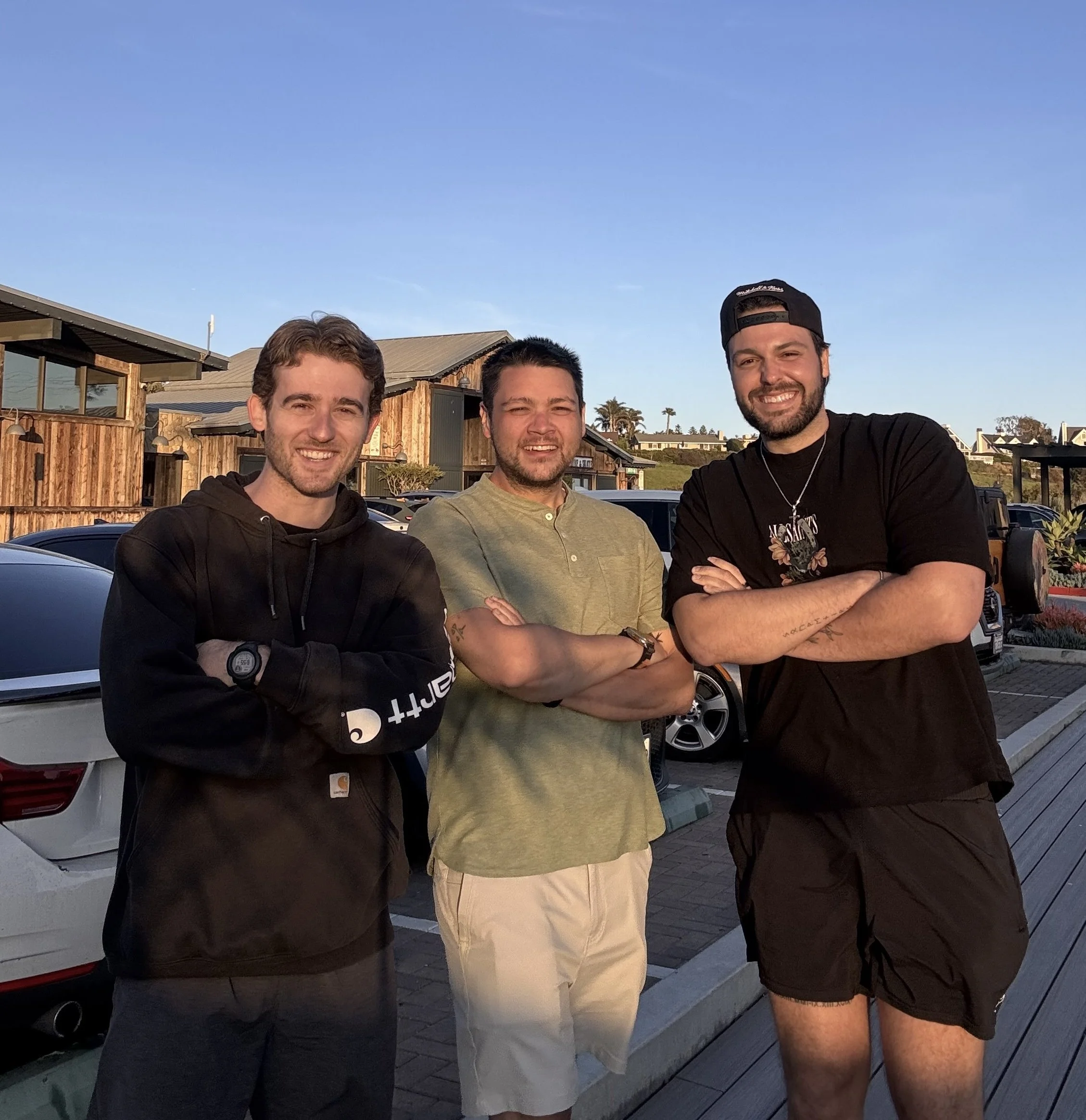 Three men standing outdoors with their arms crossed, smiling at the camera, with cars and wooden buildings in the background during sunset.