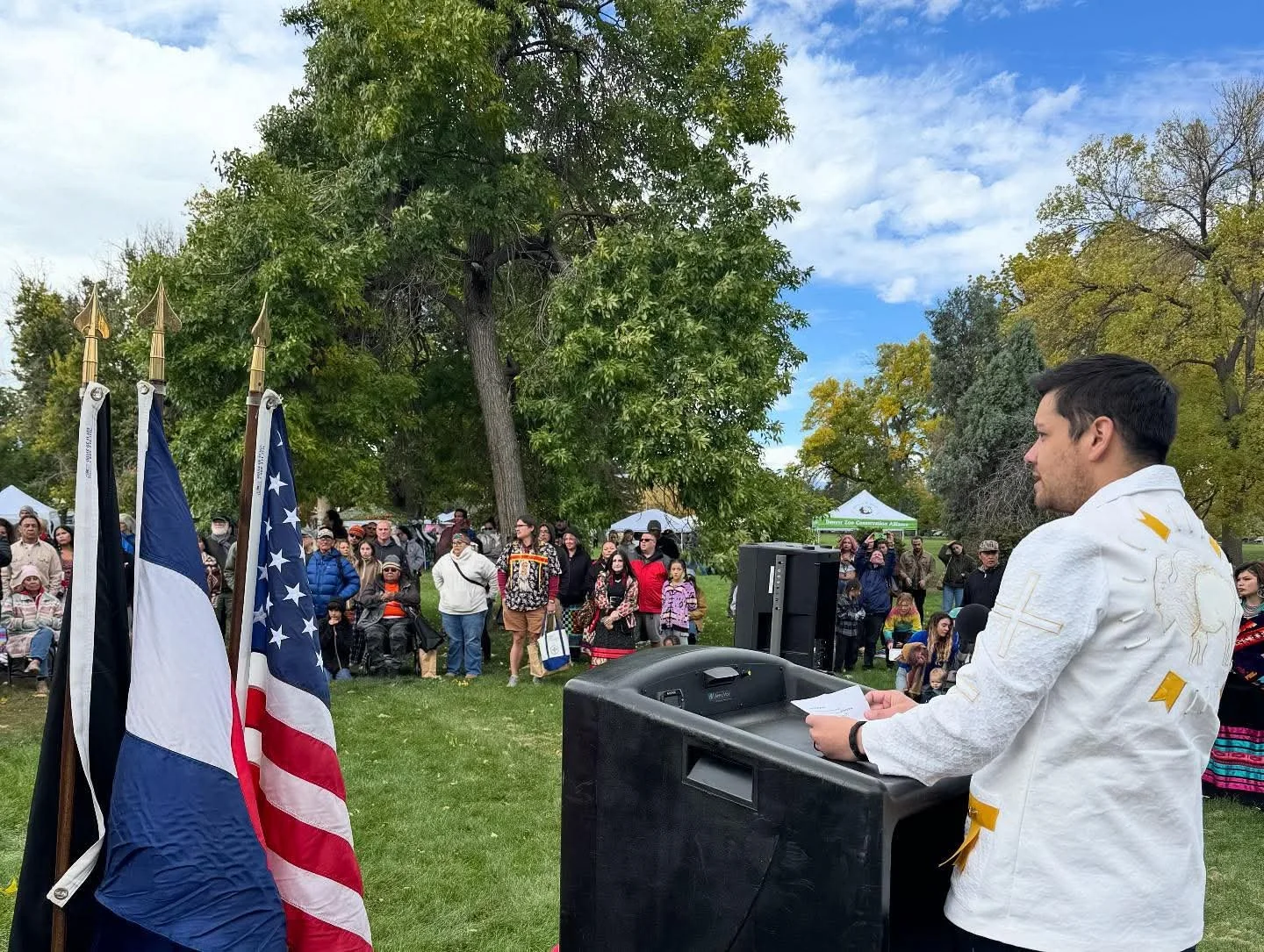 A microphone and speaker setup with an individual in white religious vestments speaking at an outdoor event, with a crowd of diverse people gathered behind, and four flags including the American flag, in a park with green trees and tents in the backg