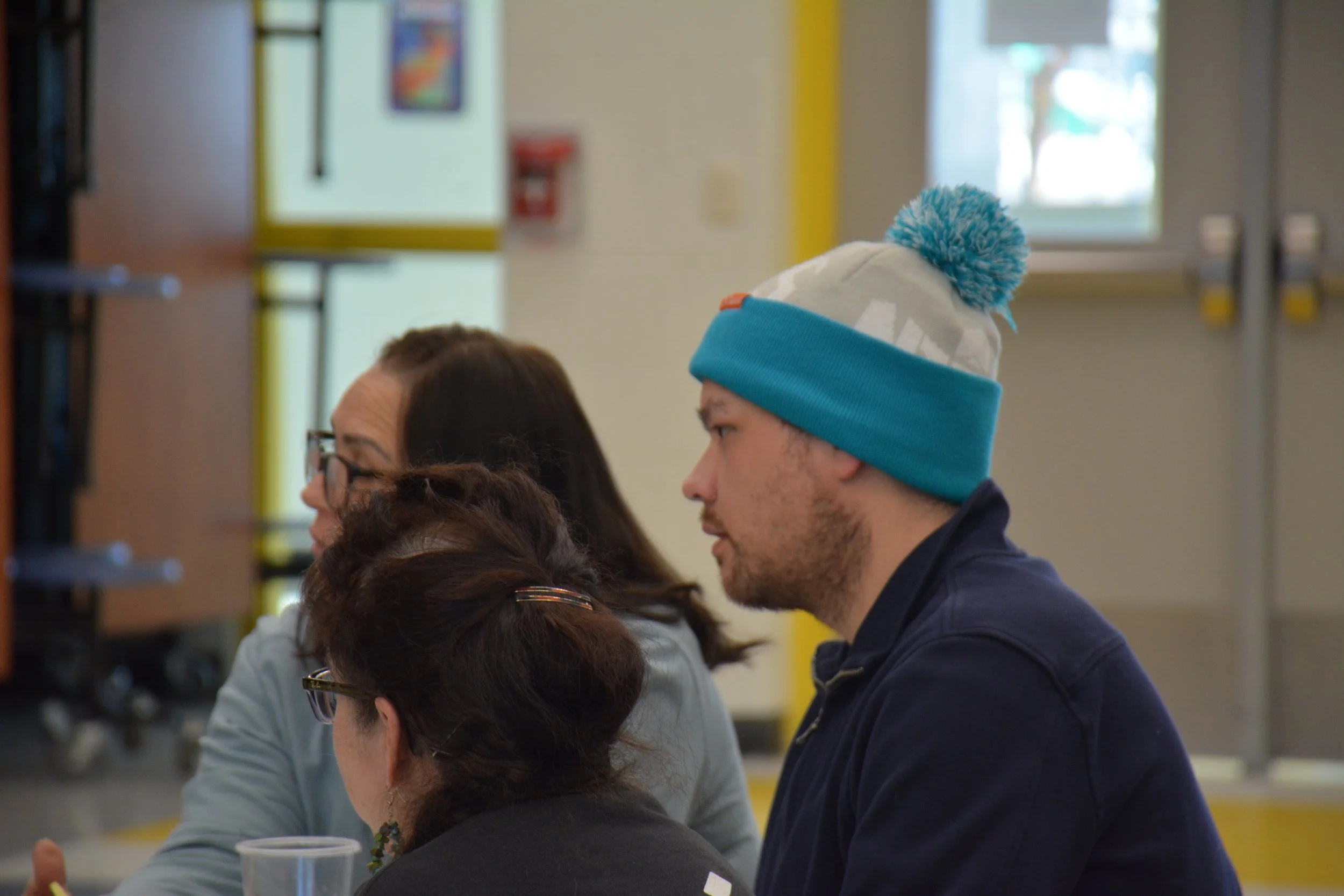 A man wearing a blue winter hat with a pom-pom, a man with glasses, a woman with glasses, and a woman with dark hair sitting indoors, possibly in a café or classroom.