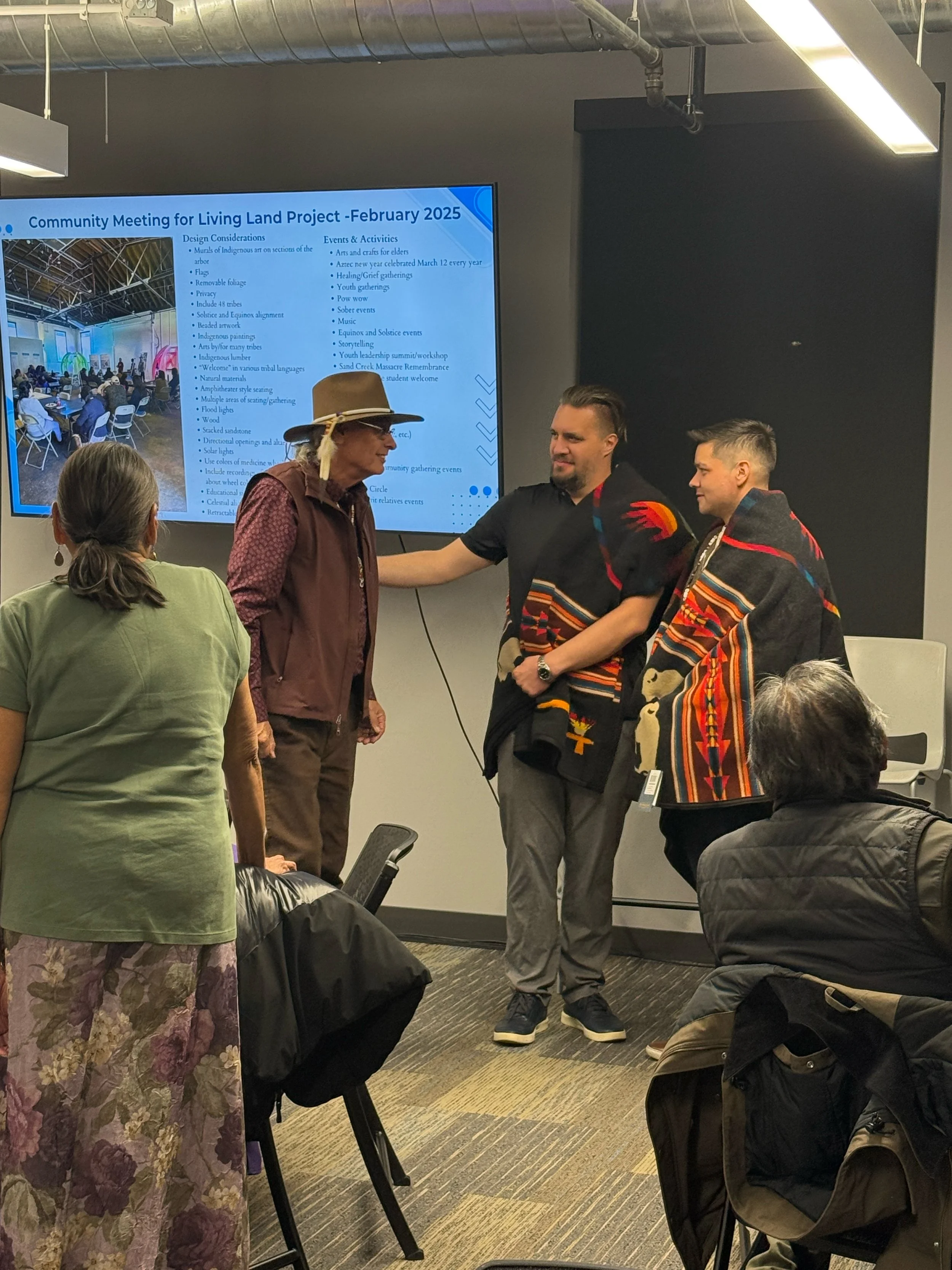 Group of people at a community meeting, two men are shaking hands, one wearing traditional Indigenous clothing with a blanket wrapped around his shoulders, and a screen behind them displays information about the event.