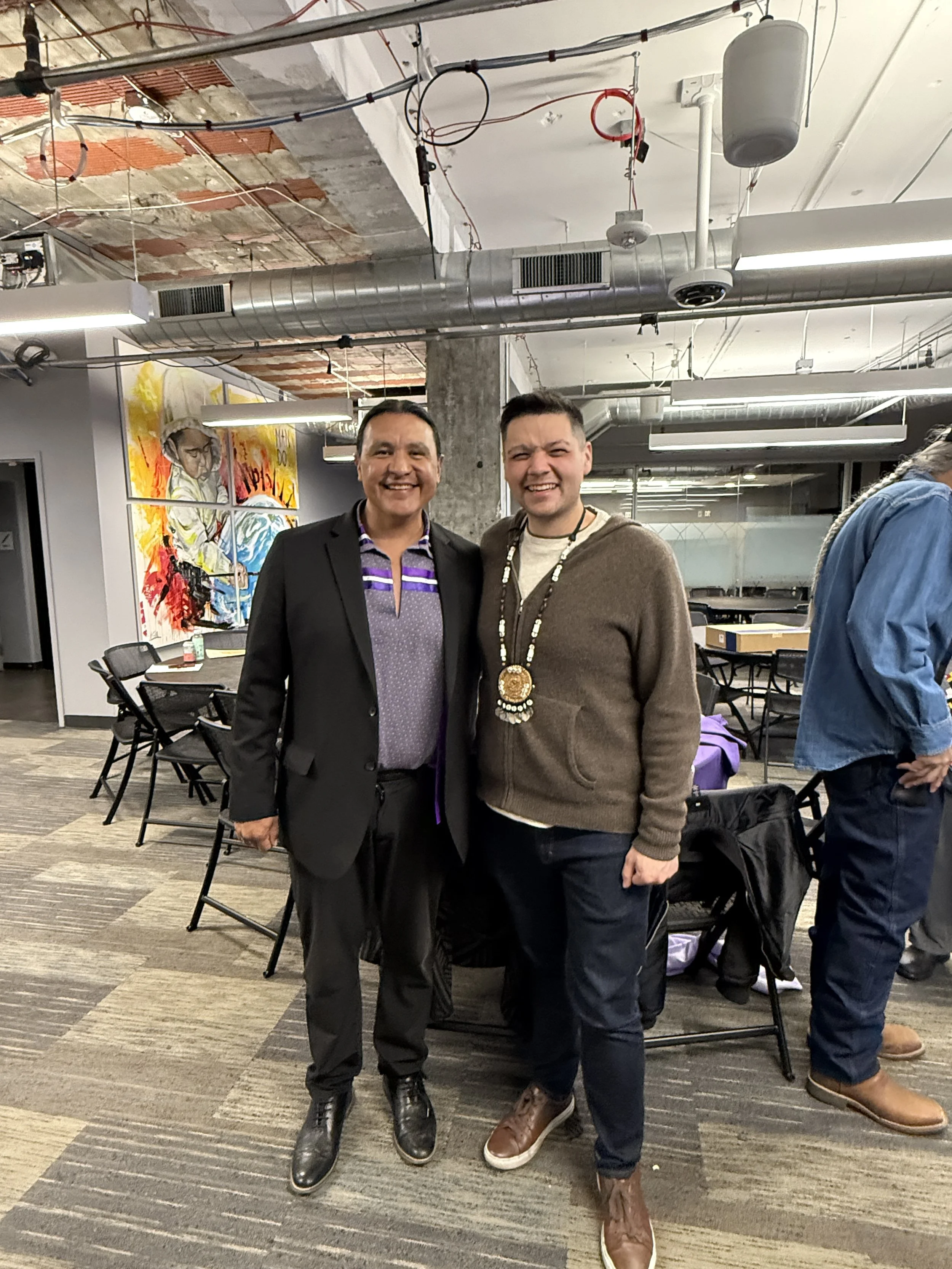 Two men standing and smiling in an office or conference room with colorful artwork on the wall behind them.
