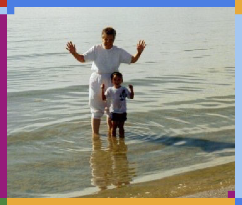 A woman and a small boy standing in shallow water at the beach, smiling and raising their arms.