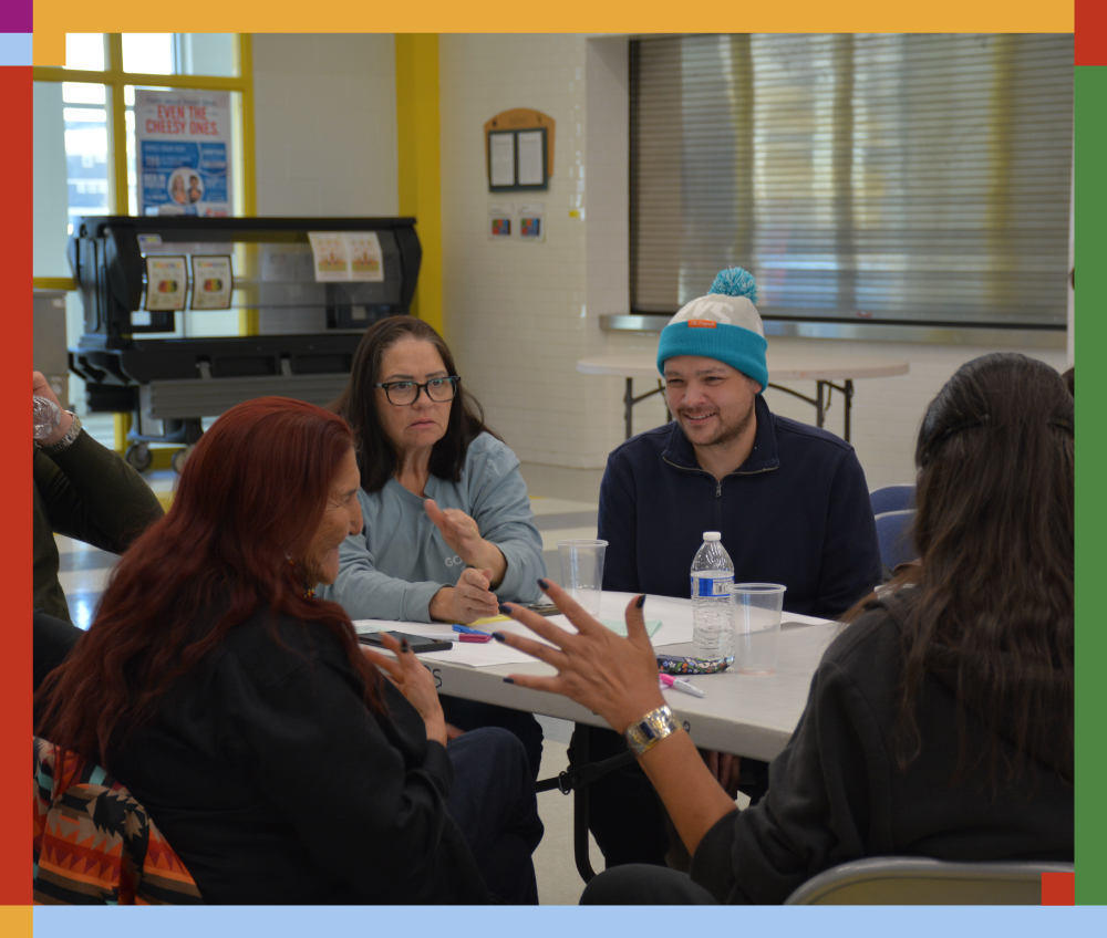 Group of people sitting around a table in a room, engaging in conversation, with some taking notes and others listening attentively.