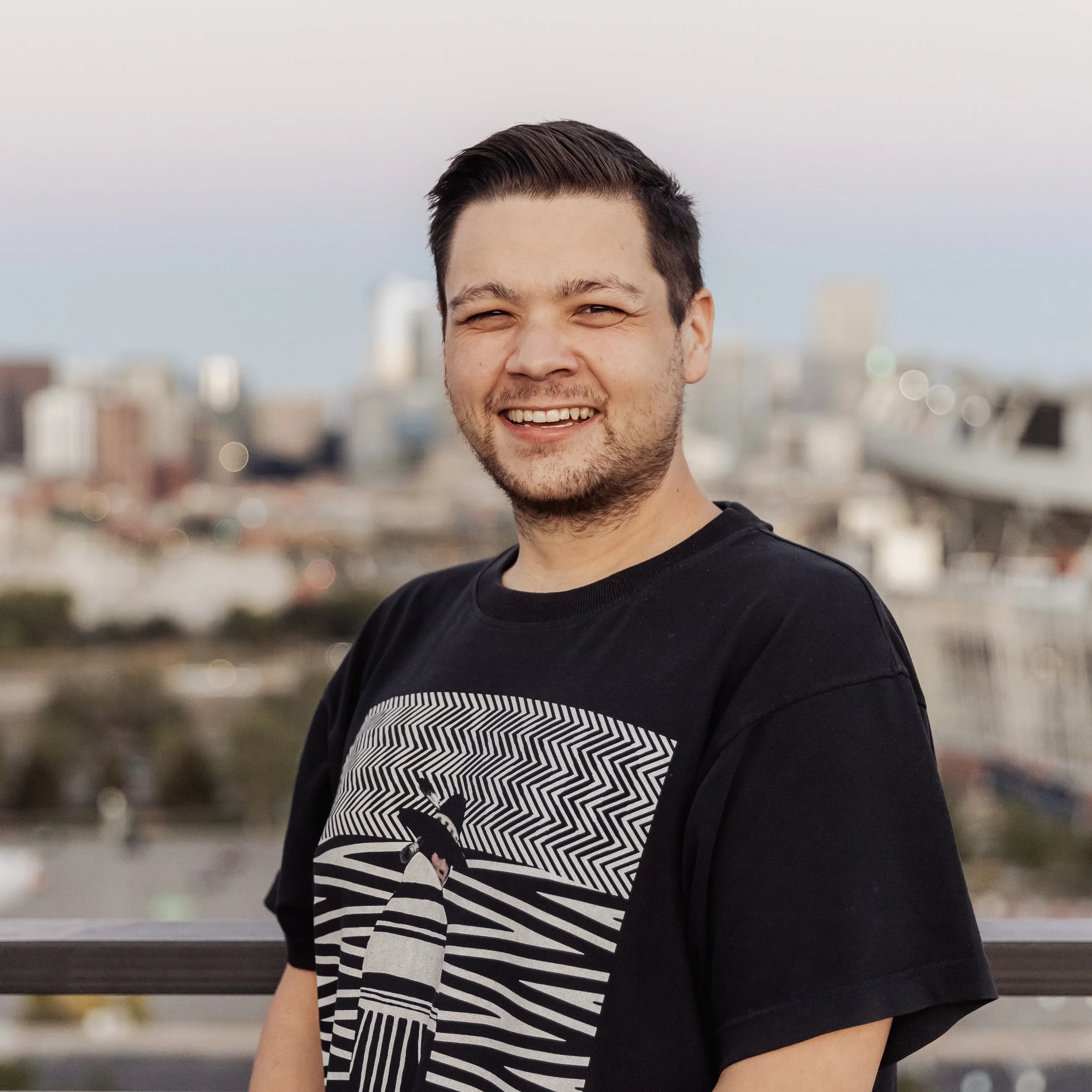 A young man with short dark hair and a beard, smiling and wearing a black graphic t-shirt, standing outdoors with a cityscape in the background during sunset.
