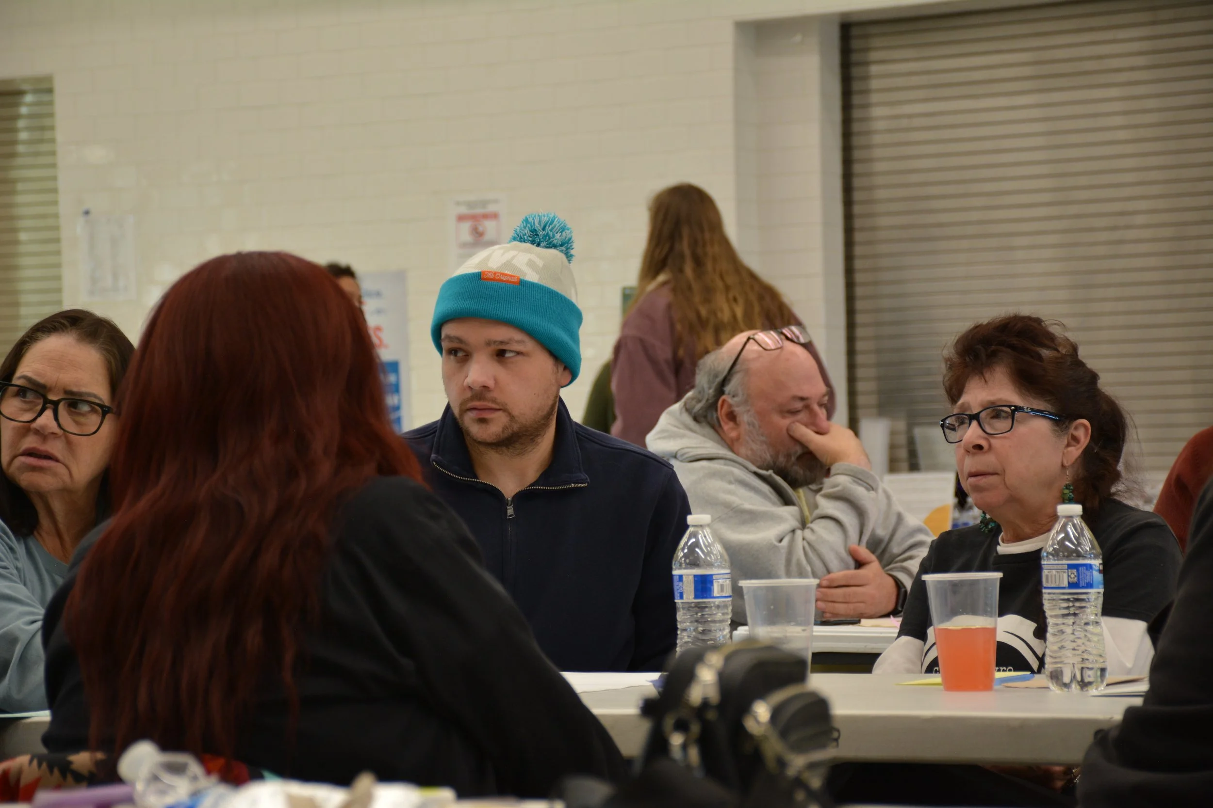Group of five people sitting at a table in a community center or cafeteria, with water bottles, cups, and papers, some wearing glasses, having a discussion.