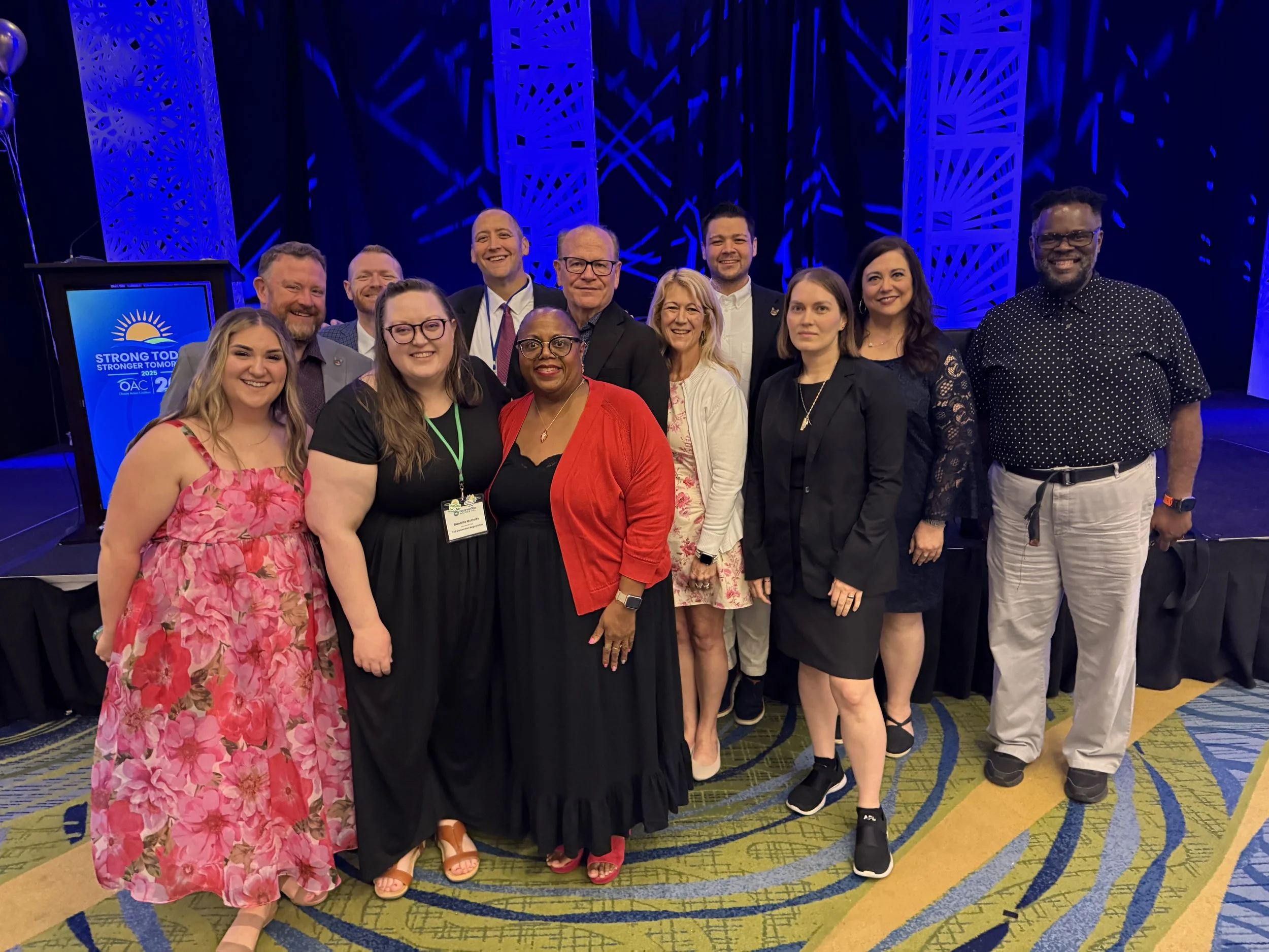 Group of diverse people smiling and posing for a photo at a conference with a blue and purple stage background and a sign that reads 'Strong Today, Stronger Tomorrow'.