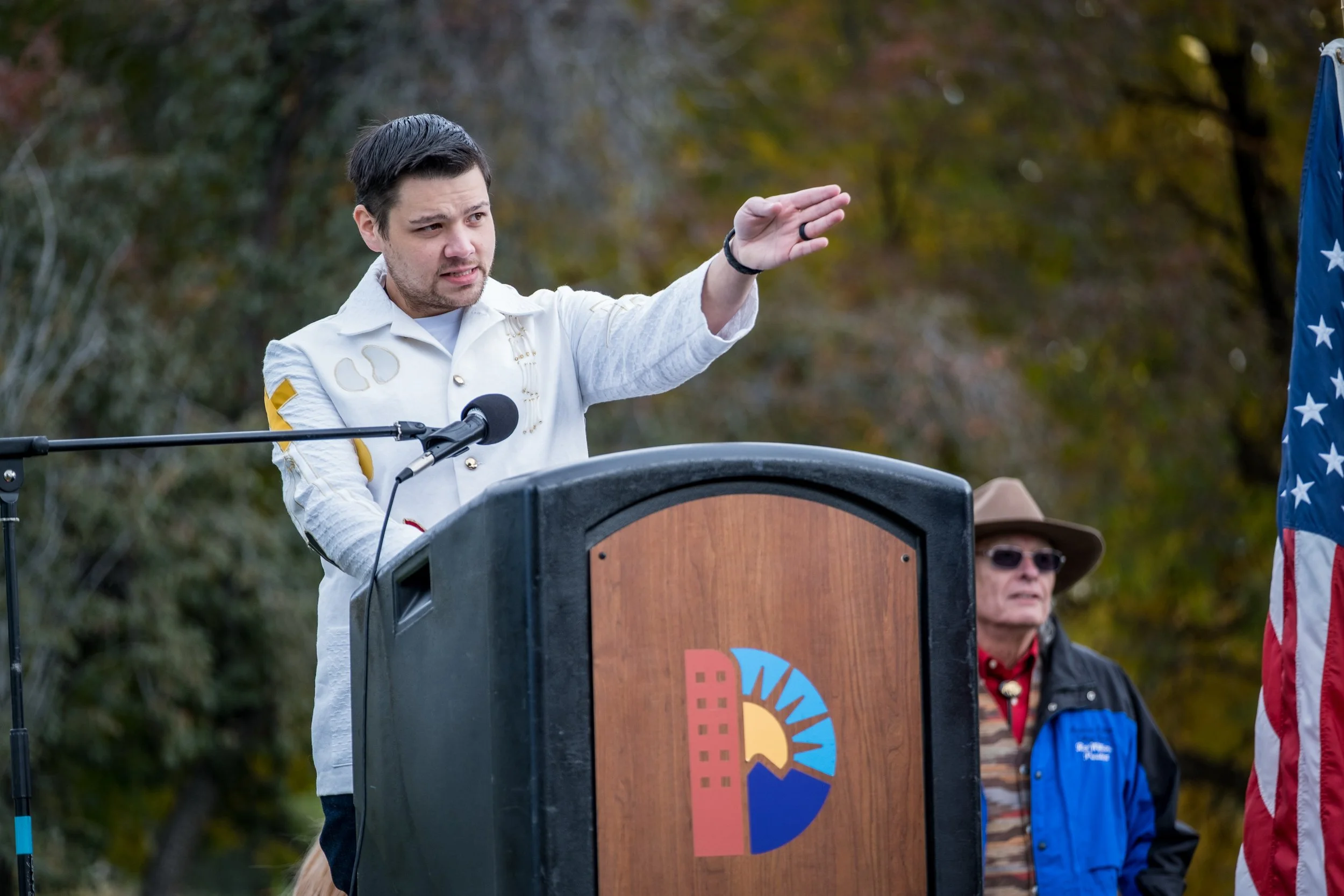 A man with dark hair and facial hair at a podium, gesturing with his right hand, wearing a white jacket with patches on the sleeves. Behind him, an older man in a wide-brimmed hat and sunglasses, dressed in a blue jacket and patterned shirt, stands n