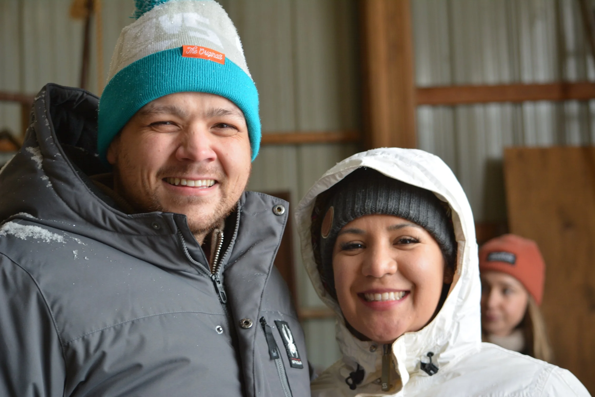 Two people smiling in winter clothing, with another person in the background, inside a wooden building.