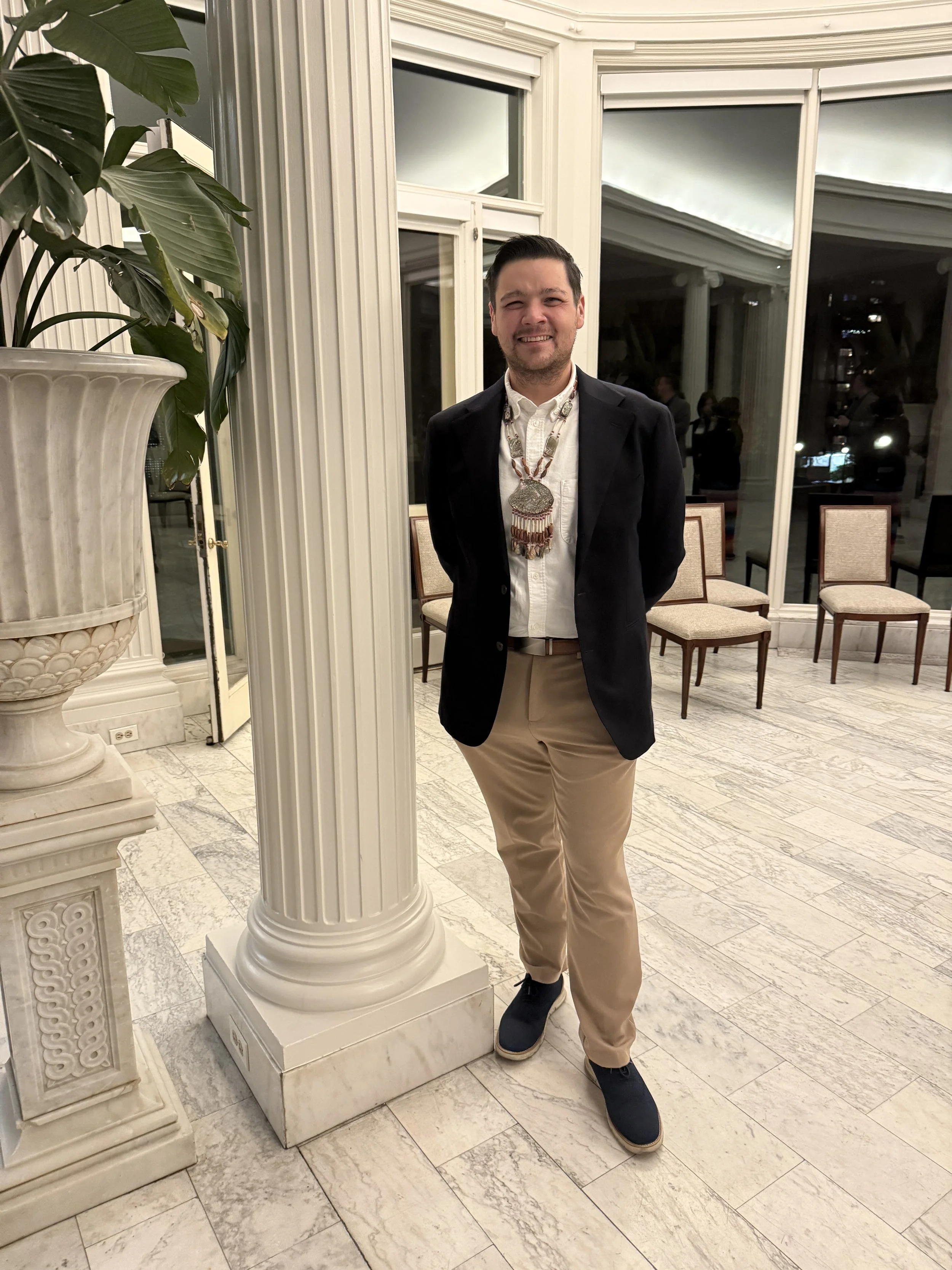 A man standing inside a room with large windows and white ornate walls, wearing a white shirt, dark blazer, beige pants, black shoes, and a large beaded necklace.