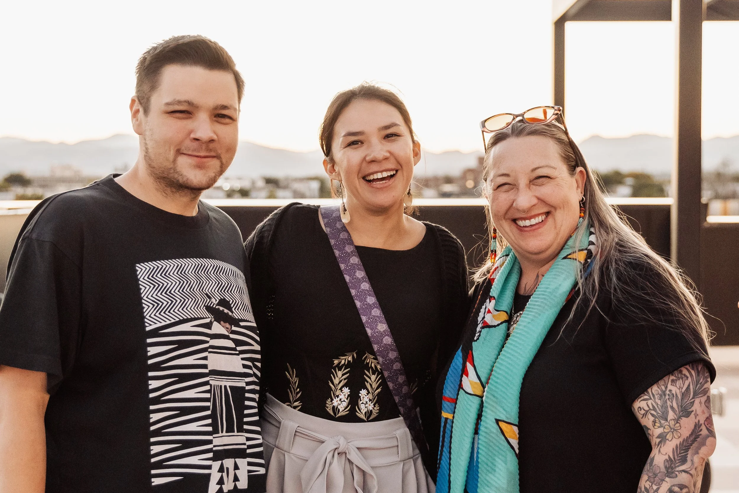 Three people smiling and standing close together outdoors, with a cityscape and hills in the background during sunset.