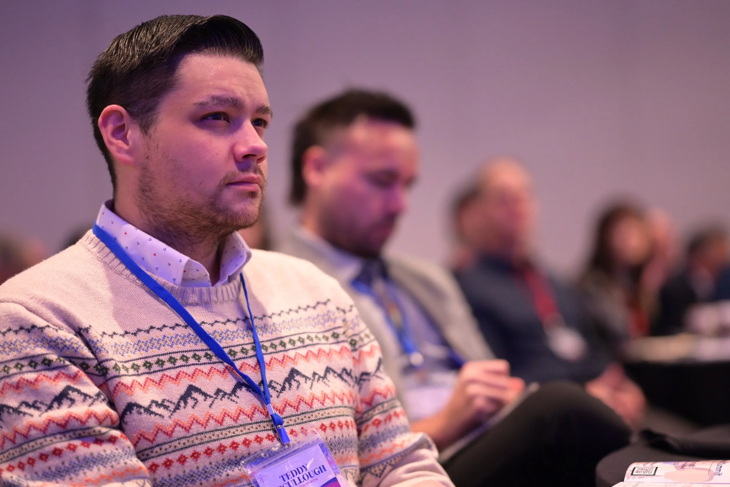 A man with dark hair and a beard wearing a tan sweater with a colorful pattern, sitting at a conference or seminar. He has a blue lanyard and a name tag that reads 'Teddy Cullough.' Other attendees are blurred in the background.