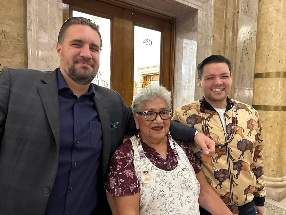 Three people standing inside a building with marble walls and a wooden door, smiling for the camera.