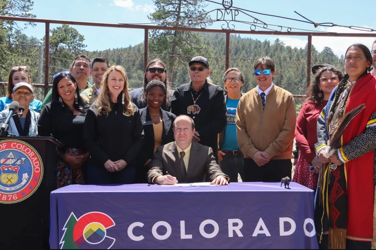 A group of people gathering outdoors with a man sitting at a table covered with a Colorado-themed cloth, surrounded by individuals of diverse backgrounds, some in traditional Native American attire, with a fence and mountainous landscape in the backg