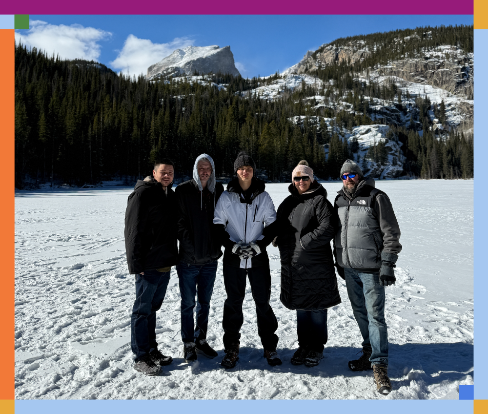 Five people standing in snow-covered landscape with mountains and evergreen trees in the background, dressed in winter clothing.