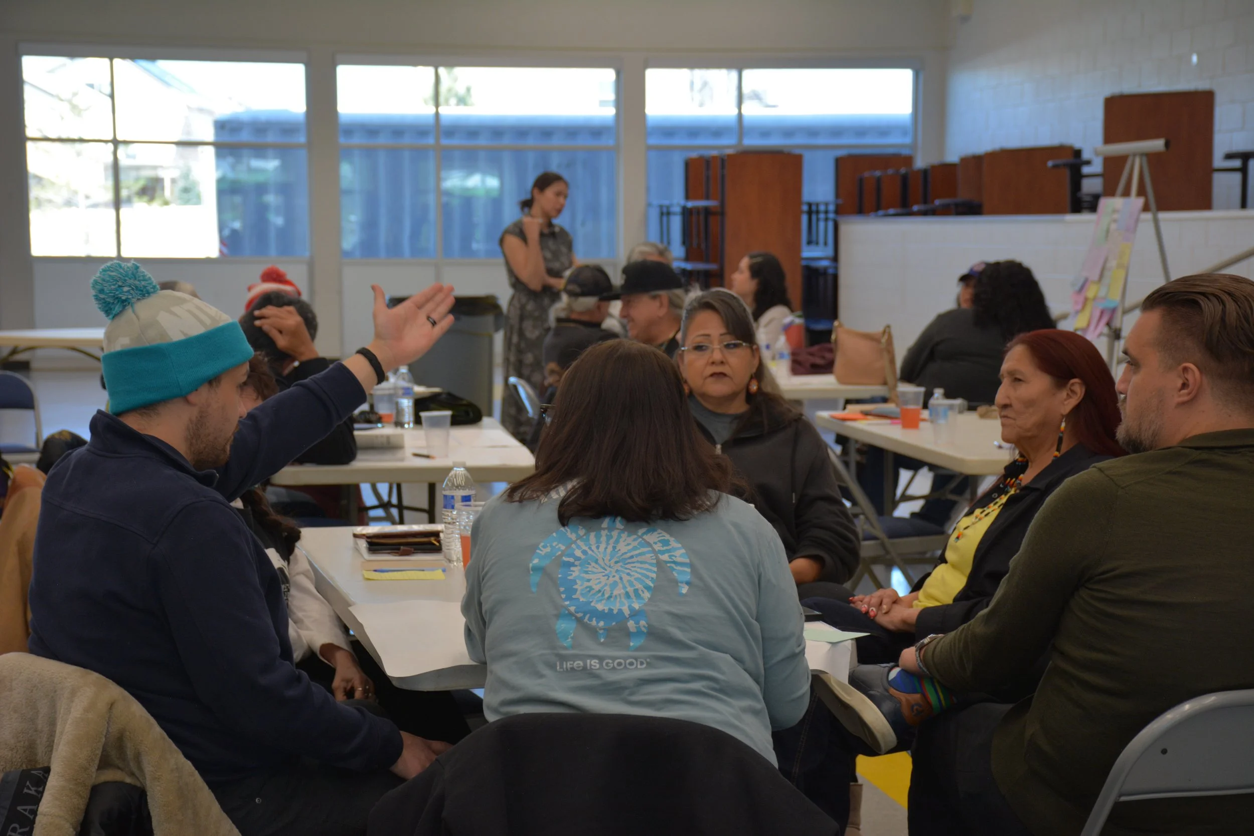 Group of people sitting at tables in a well-lit room, engaged in conversation, with a woman standing in the background.