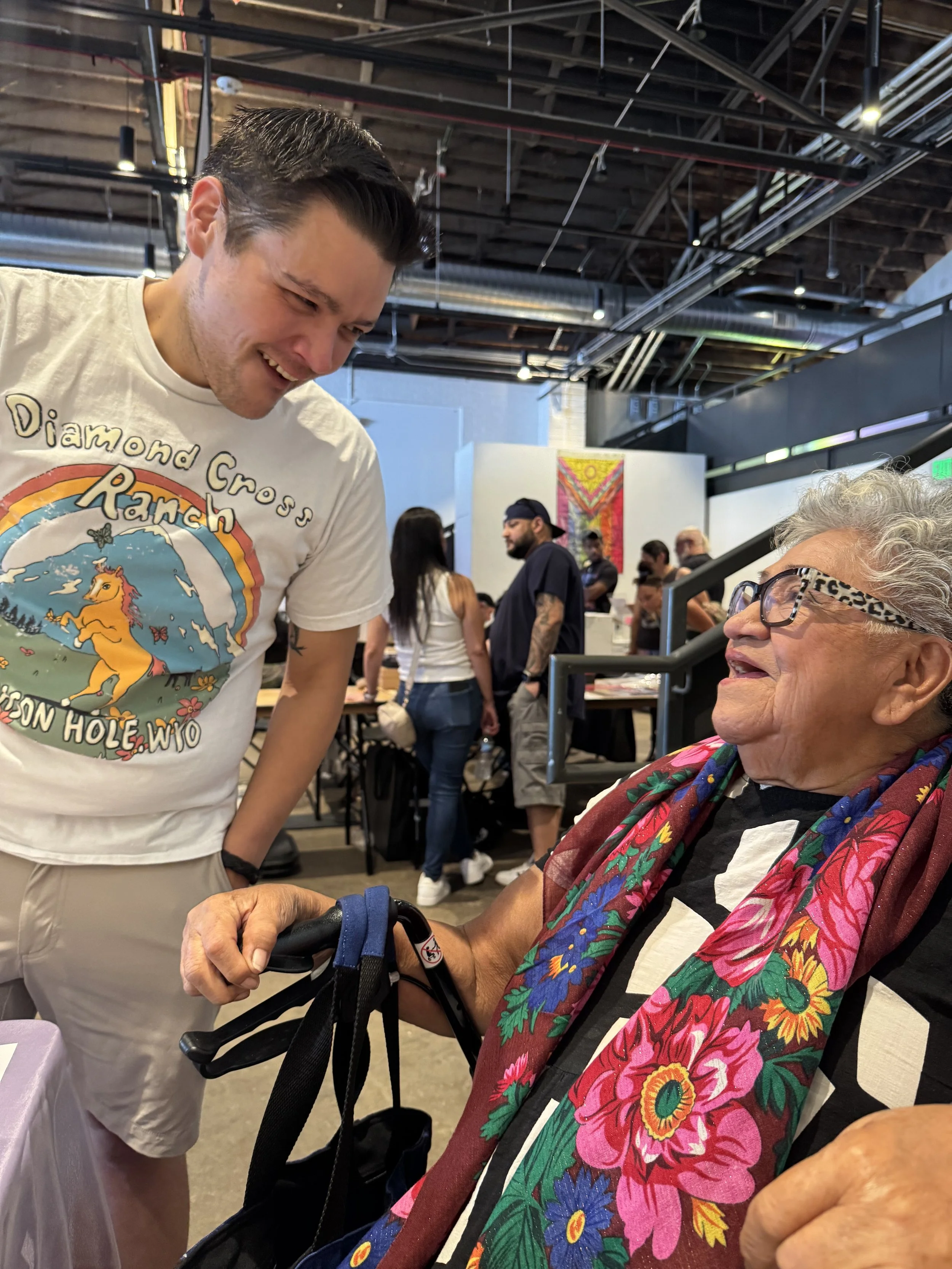 A young man with short dark hair smiling and looking down at an elderly woman with light gray curly hair, glasses, and a colorful floral scarf. The elderly woman is holding the young man's hand, which is resting on a cane. The setting appears to be a