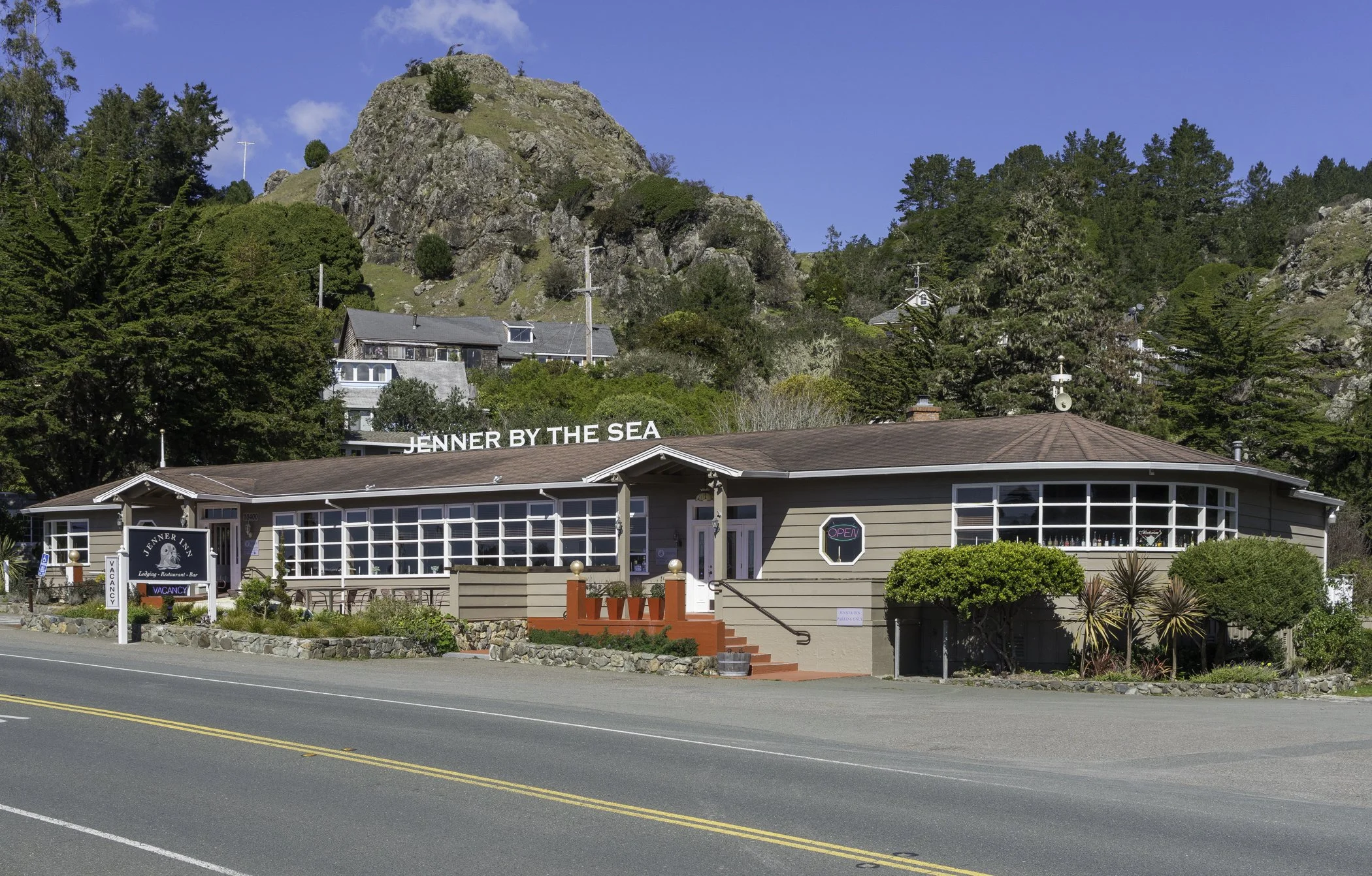 A single-story restaurant called Jenna by the Sea with a brown roof and tan siding, located along a road with a double yellow line. The restaurant has large windows, a small front porch with red stairs, and a sign that says 'Open'. There are various 