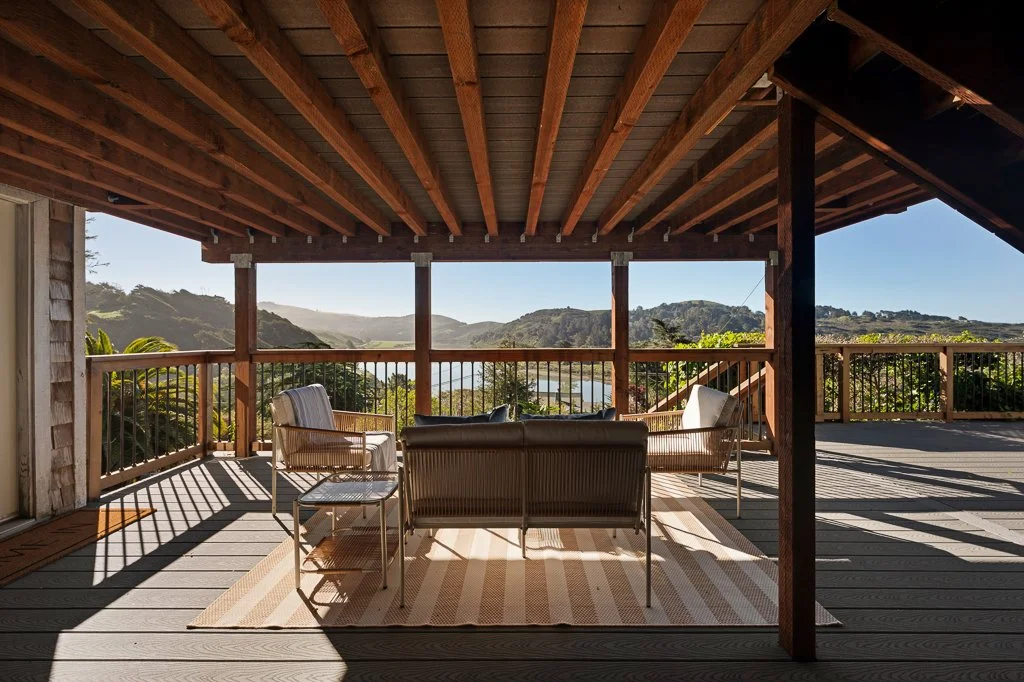 Outdoor covered patio with wooden ceiling and railing, overlooking hills and mountains, furnished with chairs, a sofa, and a striped rug.