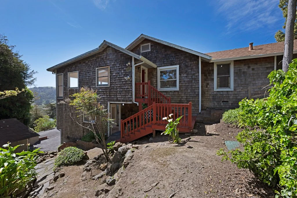 Back view of a two-story house with weathered wooden shingle siding, a red wooden staircase, and surrounding greenery on a hillside.