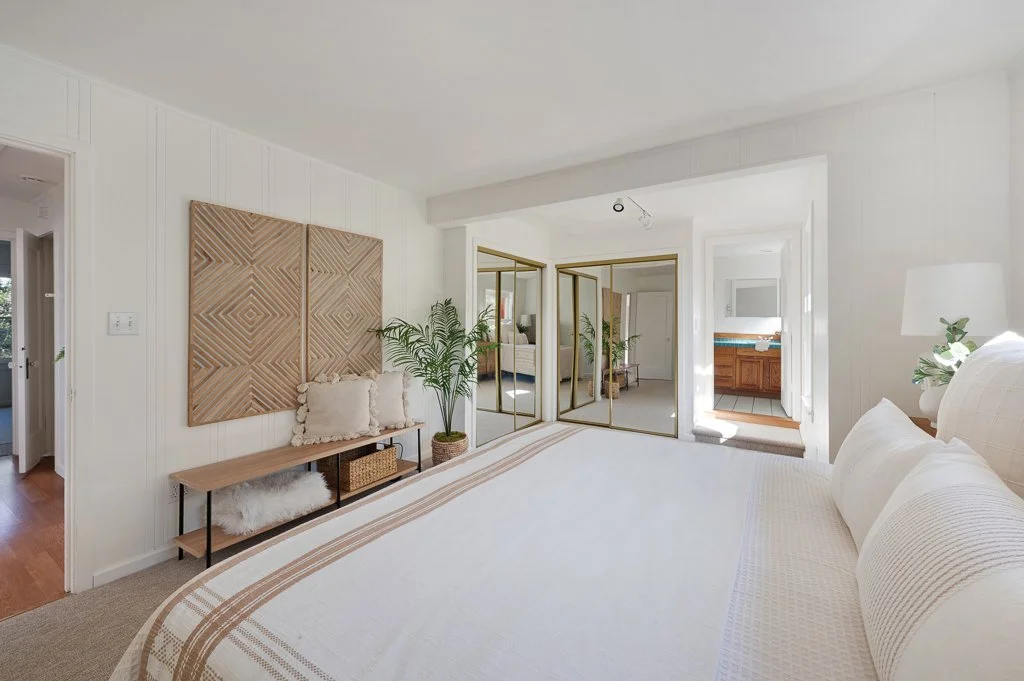 Bright white bedroom with beige accents, large bed with white bedding, wooden bench with pillows, potted greenery, mirrored closet doors, bathroom visible in the background.