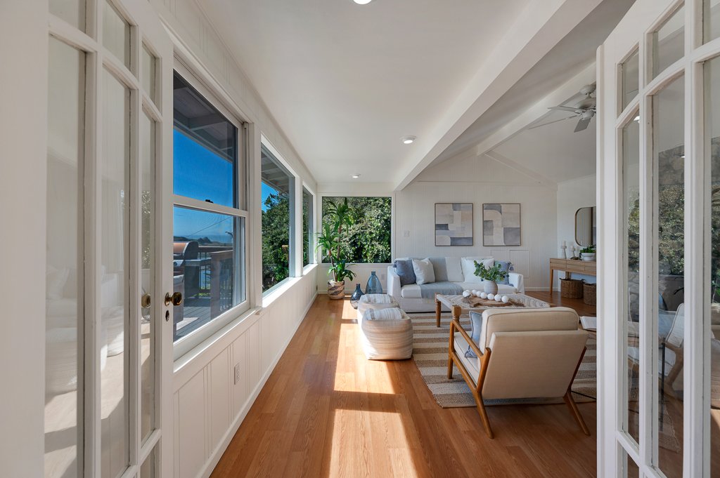 Bright living room with large windows, white sofa, beige armchair, and wooden furniture, with a view of greenery outside.