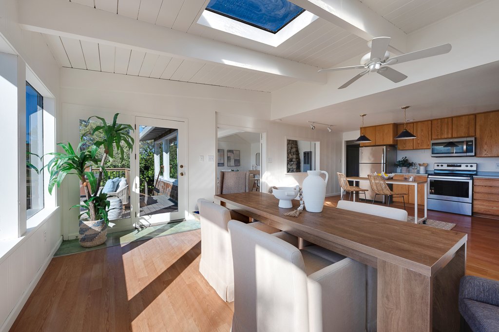 Open-concept kitchen and dining area with a large wooden table, white chairs, and decorative vases. There is a sliding glass door leading to an outdoor deck with a large potted plant and outdoor seating. The room has a white ceiling with a skylight a