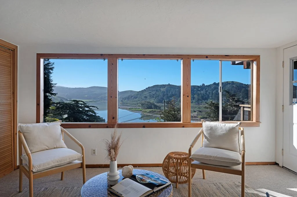 Living room with large front window showing lake and mountains, two cream armchairs, a small round table with a vase, magazine, and decorative item, beige carpet, wooden door with sliding window.
