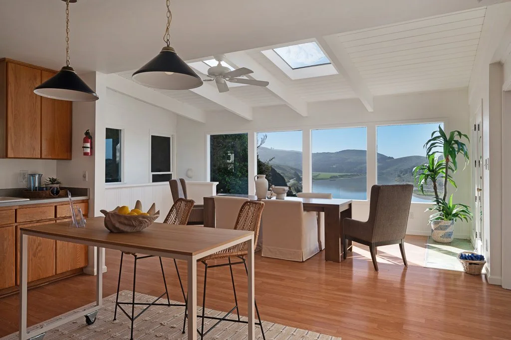 Bright living room with large windows showing a lake view, a wooden dining table with chairs, a kitchen area, and indoor plants.