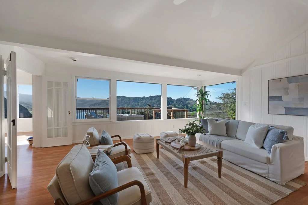 Living room with large windows showing a scenic mountain view, white walls, wood flooring, beige sofa, armchairs with light cushions, and a coffee table with decorative items.