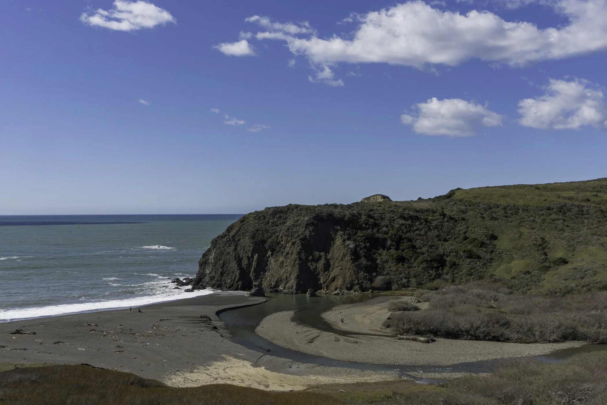 Coastal landscape with a beach, ocean, cliffs, and a stream flowing into the sea under a partly cloudy sky.