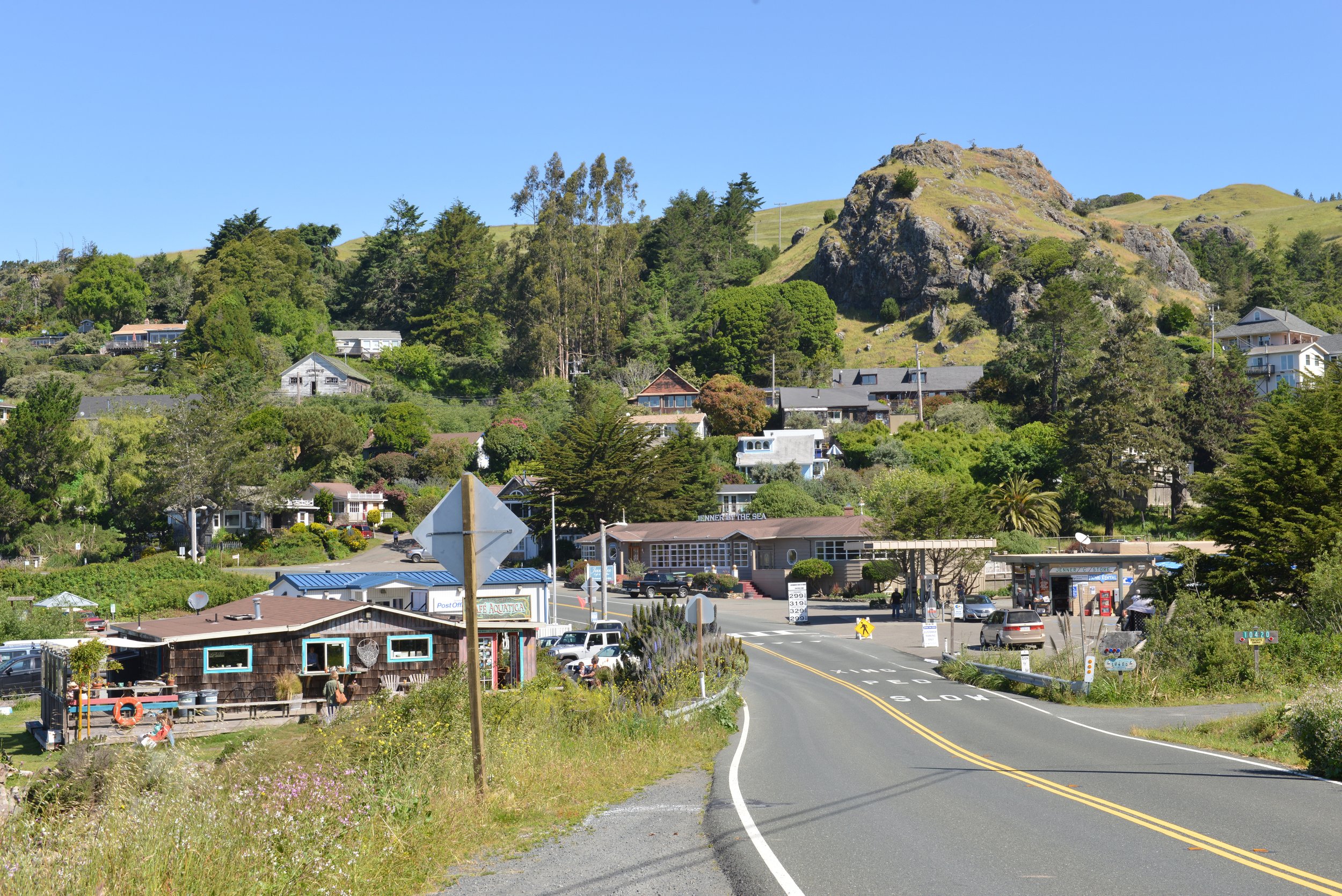 A small coastal town with houses on a hillside, surrounded by green trees and a rocky hill in the background. A road runs through the town leading to a gas station and some shops.