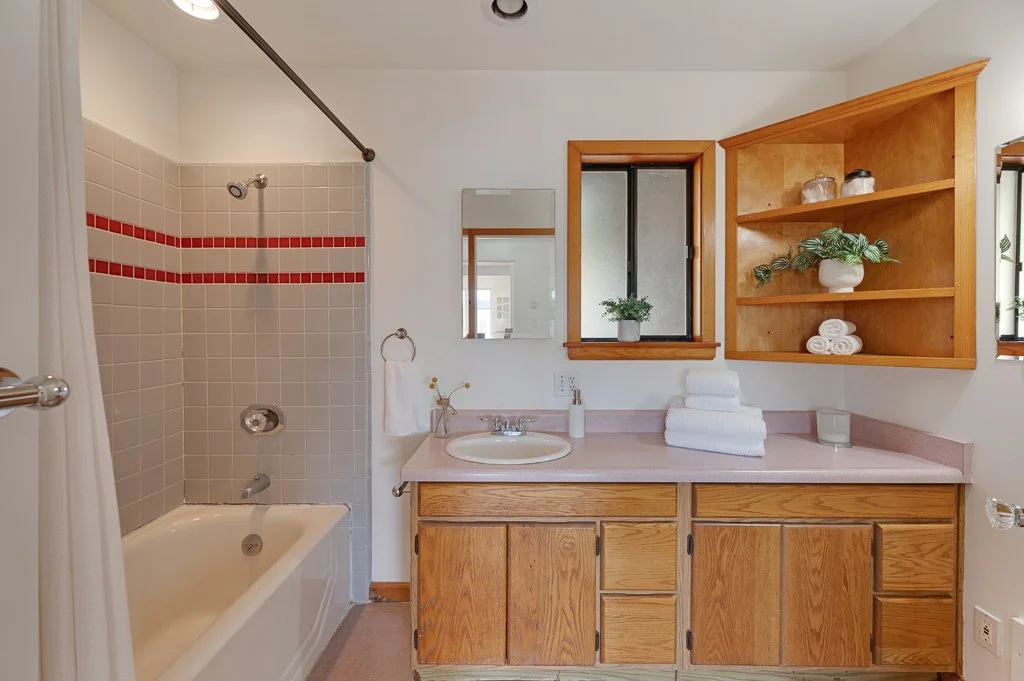 Bathroom with bathtub, shower, and wooden vanity with pink countertop, mirror, and open wooden shelves with towels and decorative items.