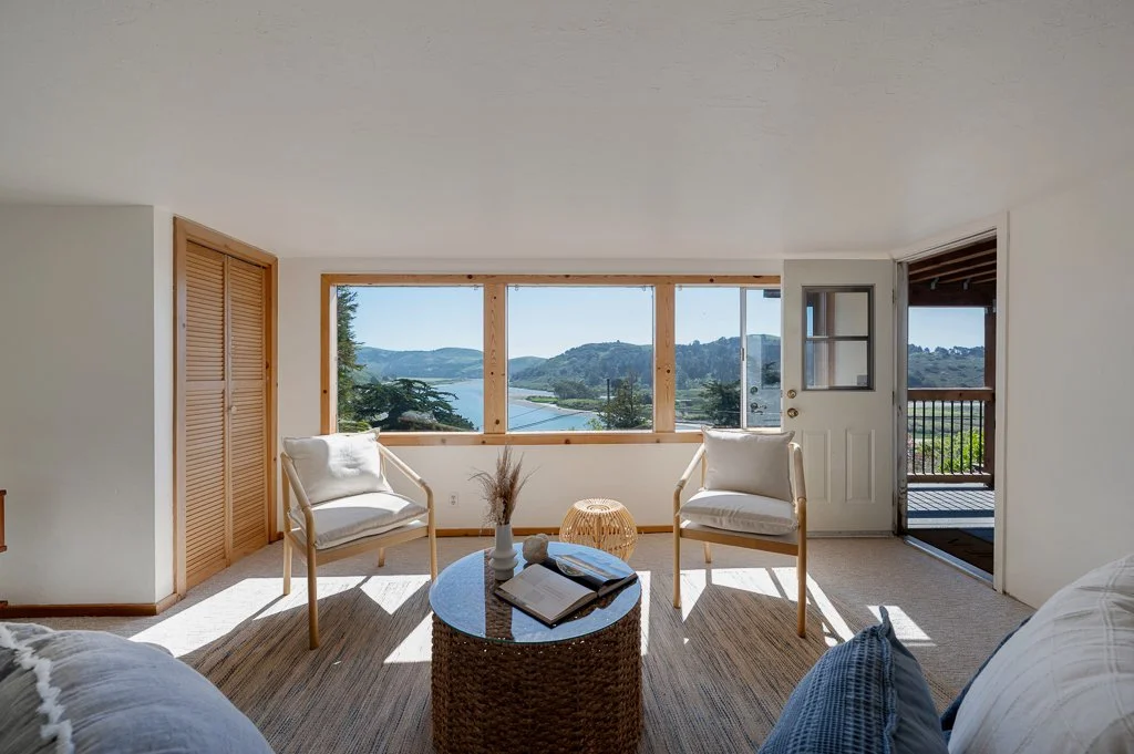 Living room with large window view of water and hills, two white armchairs, a round wicker coffee table with books and decorative items, wooden accents, and an open door leading to a balcony.