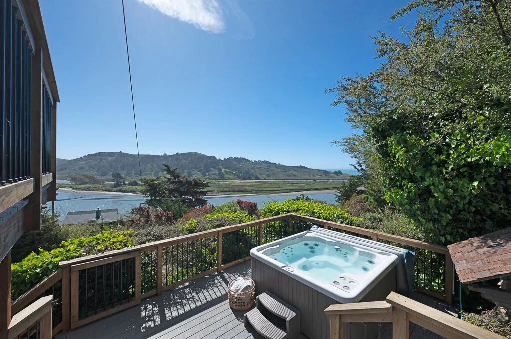 Hot tub on a wooden deck overlooking a river, surrounded by green trees and hills under a blue sky.