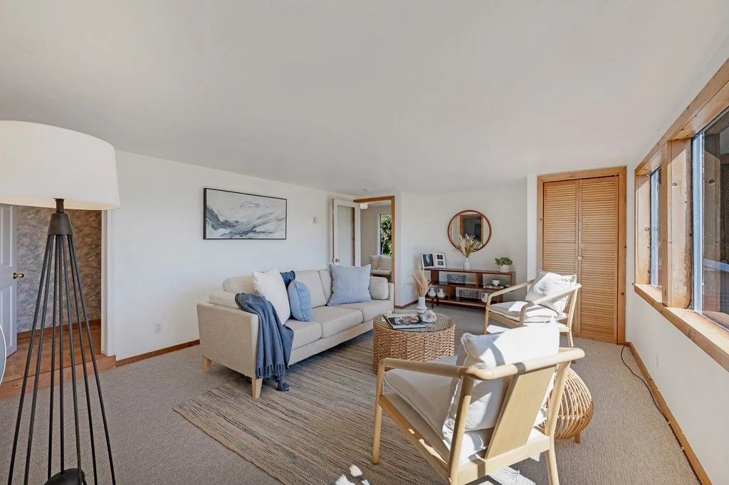 Bright living room with beige sofa, two white armchairs, and a woven coffee table. Large window with wooden trim providing natural light. Minimalist decor with a framed landscape painting and a round mirror.