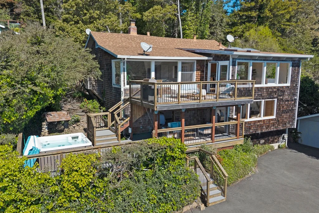 A two-story house with multiple decks, surrounded by trees, featuring a hot tub on the lower deck and satellite dishes on the roof.