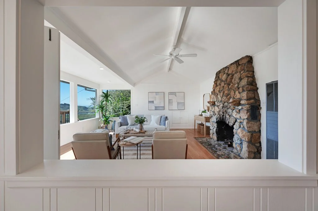 Living room with white walls, large windows, a stone fireplace, white sofa, two beige armchairs, a wooden side table, artwork on the wall, and a ceiling fan.