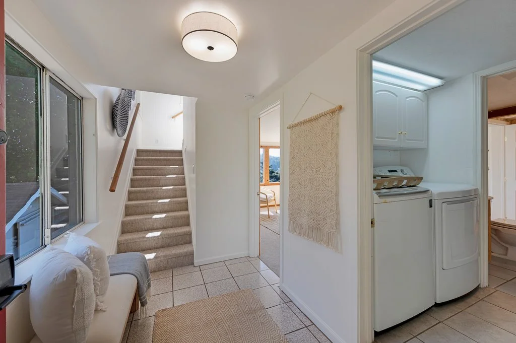 Interior view of a home featuring a stairway with carpeted steps, a laundry area with a washing machine, a window with natural light, and a hallway leading to a sitting area with a view outside.
