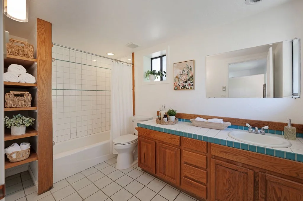 Bathroom with shower, toilet, wooden vanity with sink, mirror, window with plants, and decorative elements.