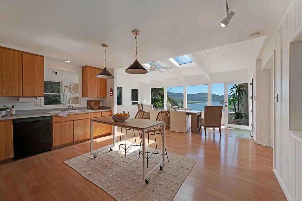 Open-concept kitchen and dining area with large windows overlooking a scenic outdoor landscape, wooden flooring, and white walls.