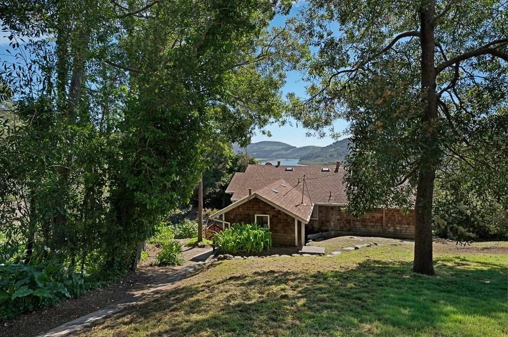 A house partially hidden behind trees, with a garden in front and hills in the background.