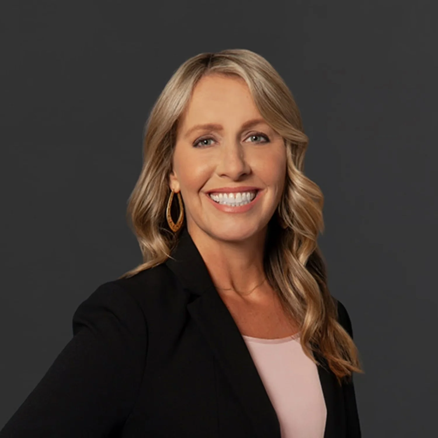 A woman with blonde hair styled in loose waves, wearing a black blazer over a light pink top, smiling against a dark gray background.