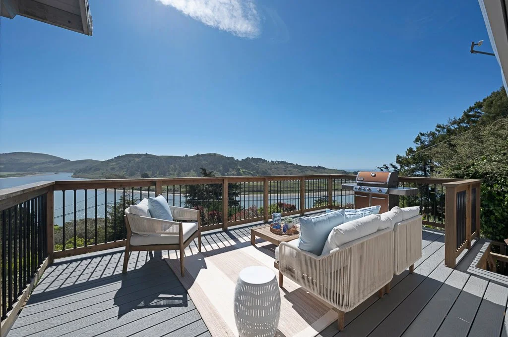 A balcony with outdoor furniture including a sofa, chair, and rug, overlooking a body of water and green hills under a clear blue sky.